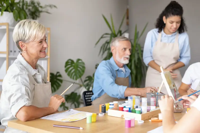 Group of three seniors engaging in a creative painting workshop, smiling and surrounded by colorful paint supplies and art materials in a bright, plant-filled studio.