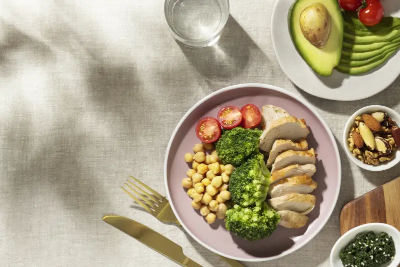 Healthy meal featuring sliced chicken breast, chickpeas, steamed broccoli, and cherry tomatoes, served in a pink bowl with a side of avocado and a glass of water on a light tablecloth.