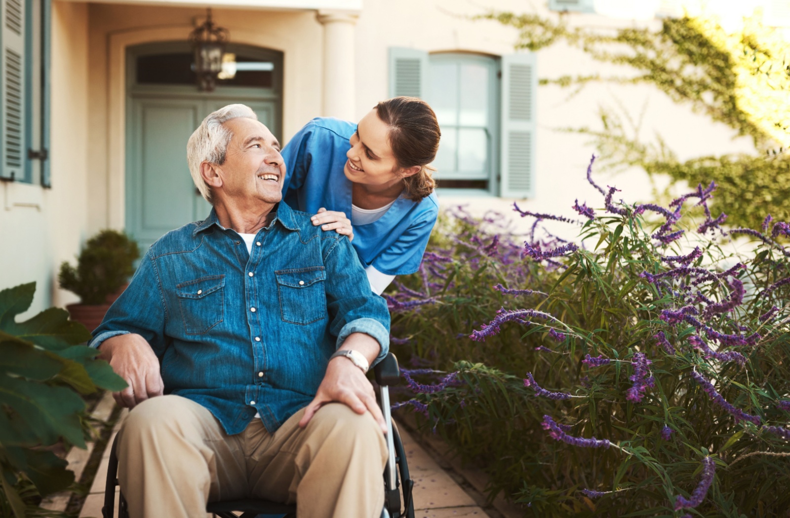 A nurse smiles at a senior in a wheelchair while providing them a hand at getting around their assisted living home
