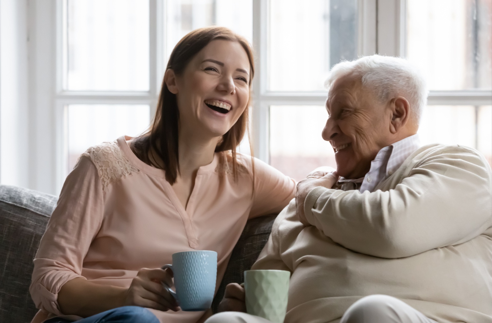 An adult and senior parent sitting on a sofa laughing in conversation while drinking coffee
