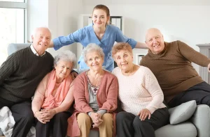Group of six smiling seniors with a caregiver in a cozy living room, promoting companionship and elder care.