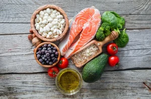 Fresh ingredients for a healthy meal, including salmon fillets, spinach, tomatoes, avocado, blueberries, quinoa, and olive oil, arranged on a rustic wooden surface.