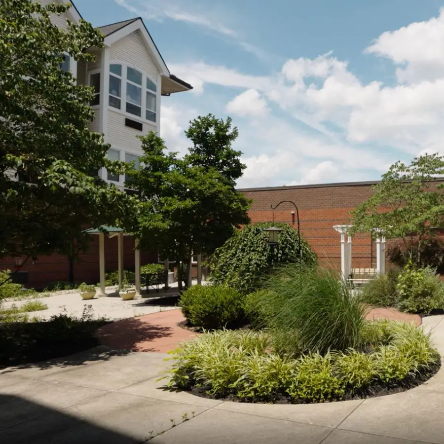 Lush courtyard with greenery and landscaping, featuring a circular flower bed, trees, and a brick building under a blue sky with clouds.
