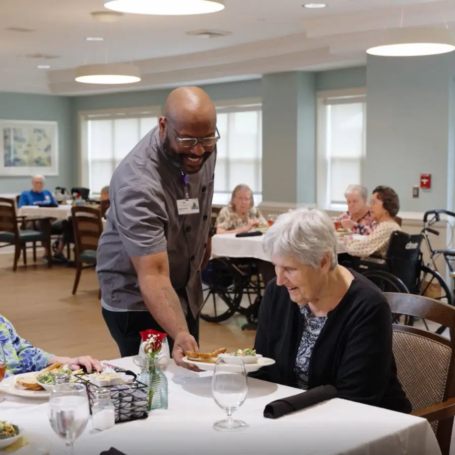 A caregiver serves a meal to an elderly woman in a dining area, with other residents enjoying their meals in the background. The setting is bright and welcoming, highlighting a community atmosphere in a senior living facility.