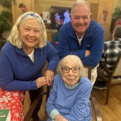 Three smiling individuals, including an elderly woman in a blue sweater seated in a chair, flanked by two adults in matching blue attire, posing together in a cozy indoor setting with festive decorations and a TV in the background.