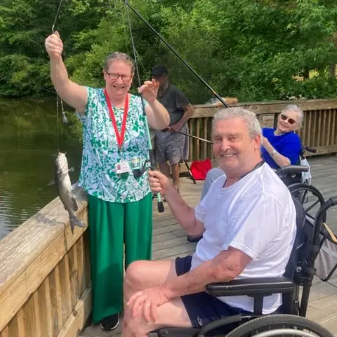 A group of people enjoying a fishing trip at a lake, with a woman holding up a caught fish while a man in a wheelchair smiles nearby. Other individuals are seated in the background, highlighting a relaxed outdoor activity.