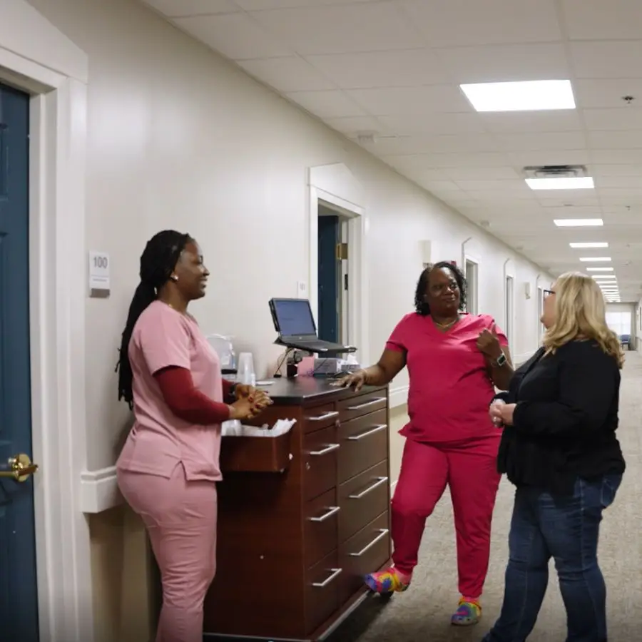 Healthcare professionals interacting in a hospital corridor, with one nurse at a reception desk and two women discussing nearby, showcasing a collaborative medical environment.