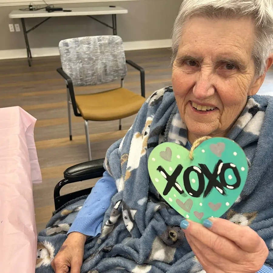 Elderly woman in a cozy blanket smiles while holding a painted heart with "XOXO" in a community activity setting.