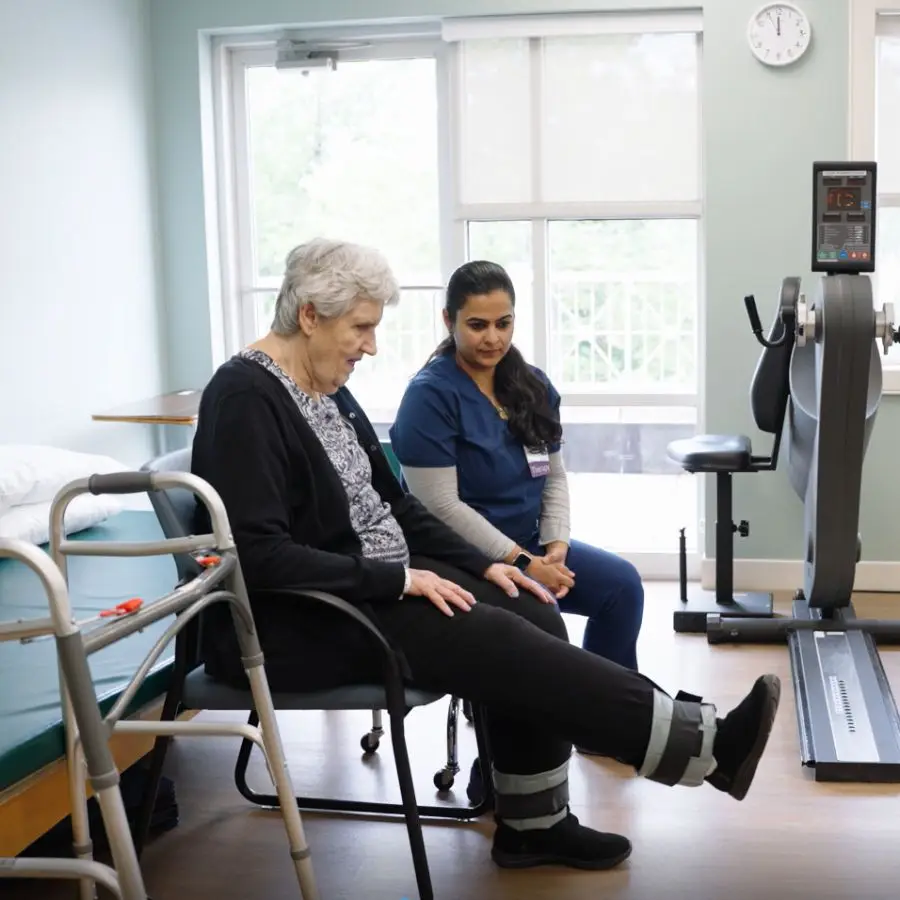 Senior woman receiving physical therapy assistance from a healthcare professional in a rehabilitation setting. The therapist guides the patient in leg exercises while seated, promoting mobility and recovery.