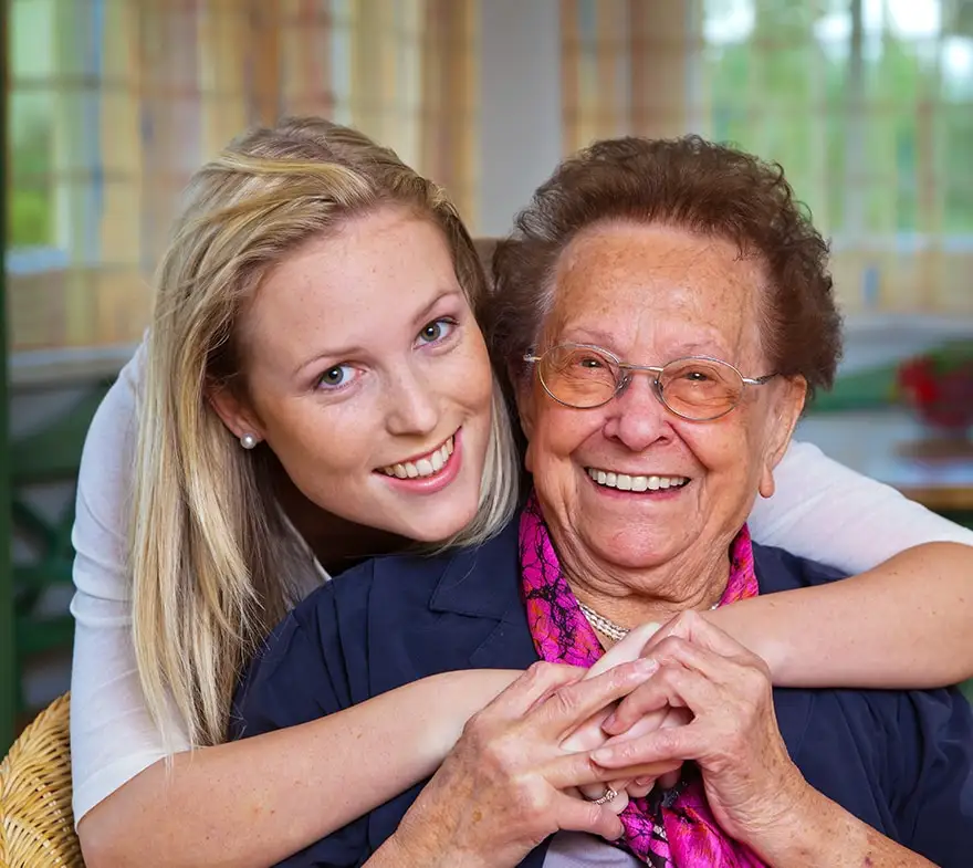 Young woman embracing and smiling with elderly woman, both enjoying a joyful moment together in a bright indoor setting.