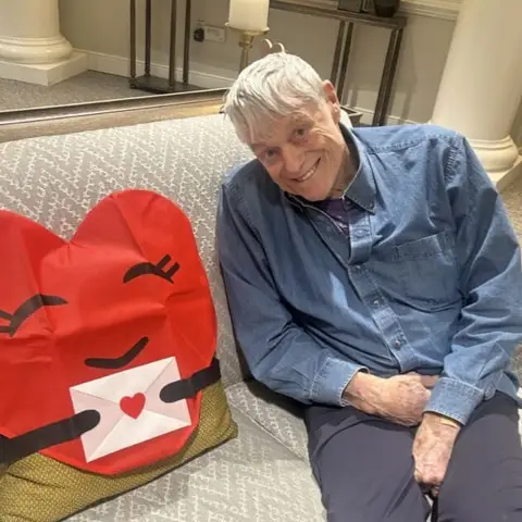 An elderly man sitting on a couch next to a large, heart-shaped pillow with a smiling face, creating a warm and inviting atmosphere.