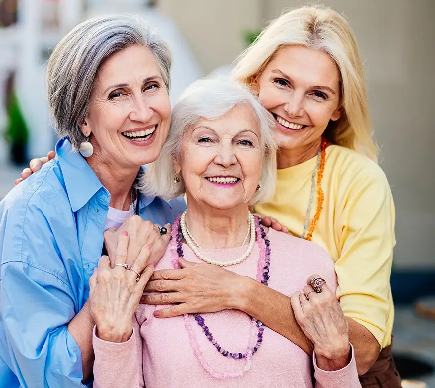 Three smiling women of different ages share a joyful moment, showcasing a strong bond of friendship and family. The group includes a senior woman in a pink sweater, flanked by two middle-aged women in blue and yellow tops, all standing outdoors.