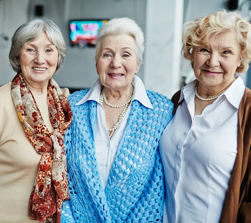 Three smiling senior women standing together in a bright indoor setting, showcasing friendship and joy. They are dressed in casual attire, with one wearing a blue cardigan and another with a colorful scarf.